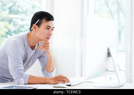 Pensive young Vietnamese ui designer with pencil behind his ear looking at computer screen Stock Photo