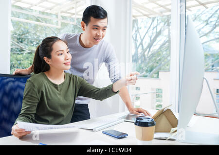 Smiling young Asian website designers discussing interface on computer screen at meeting Stock Photo