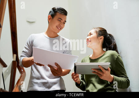Cheerful Vietnamese coworker with tablet computer and documents discussing last corrections of design project Stock Photo
