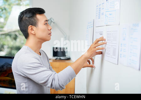 Pensive young Asian UX designer looking at documents with wireframes hanging on wall in office Stock Photo