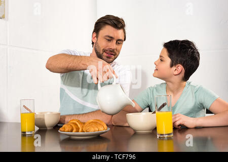 smiling dad pouring milk to son during breakfast in kitchen Stock Photo