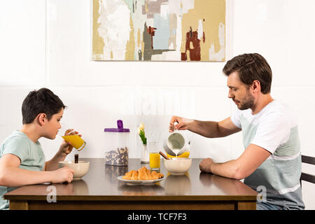 father pouring milk in bowl while sin drinking orange juice during breakfast Stock Photo
