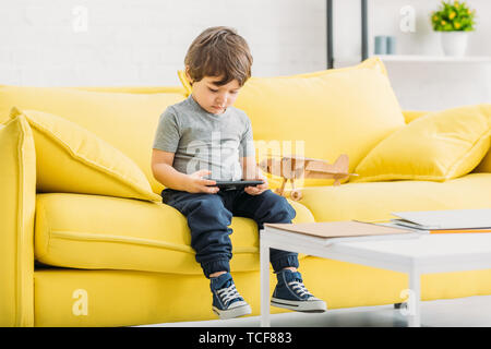 cute boy sitting on yellow sofa near wooden plane model and holding digital tablet Stock Photo
