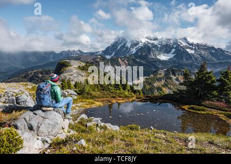 Female hiker resting on a rock at a small mountain lake, view from Tabletop Mountain to Mt. Shuksan with snow and glacier, Mt. Baker-Snoqualmie Nation Stock Photo