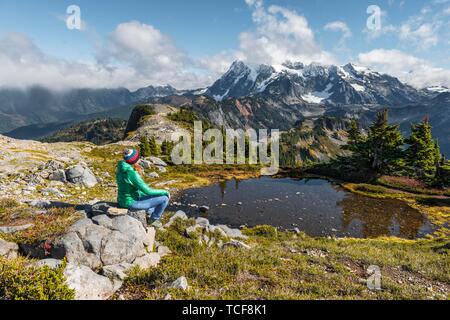 Female hiker resting on a rock at a small mountain lake, view from Tabletop Mountain to Mt. Shuksan with snow and glacier, Mt. Baker-Snoqualmie Nation Stock Photo