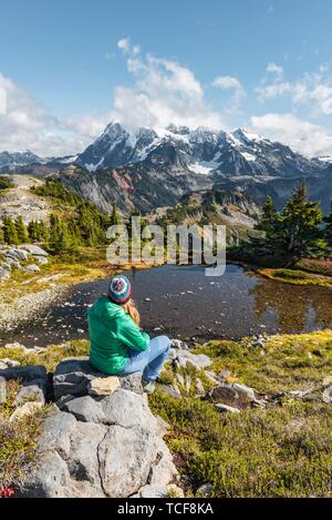 Female hiker resting on a rock at a small mountain lake, view from Tabletop Mountain to Mt. Shuksan with snow and glacier, Mt. Baker-Snoqualmie Nation Stock Photo