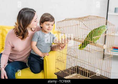 happy mother with adorable son looking at green parrot in bird cage at home Stock Photo