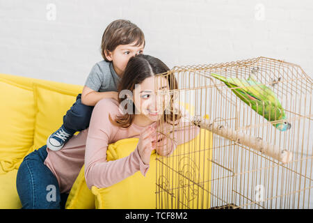 smiling mother and son looking at green parrot in bird cage at home Stock Photo