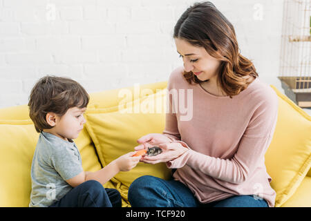 smiling mother with adorable son holding cute hamster while sitting on yellow sofa Stock Photo