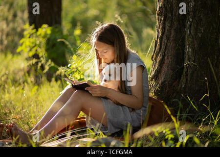 A girl sitting near a tree and reading a book, holding a labrador puppy. At sunset in the forest in summer. The concept of friendship, happiness, joy  Stock Photo