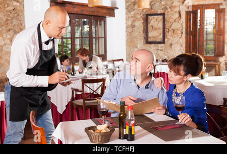 Cheerful waiter taking order of happy man and woman having dinner at cafe Stock Photo
