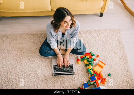 overhead view of smiling woman sitting on carpet near toy blocks and using laptop Stock Photo