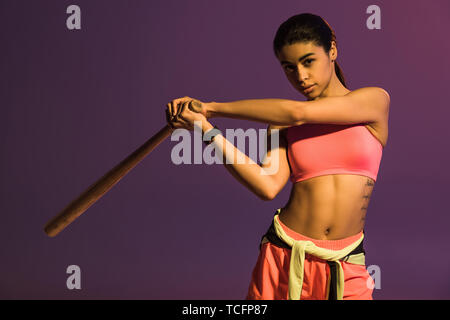 sportive african american girl in pink sports bra holding baseball bat and looking at camera isolated on purple Stock Photo