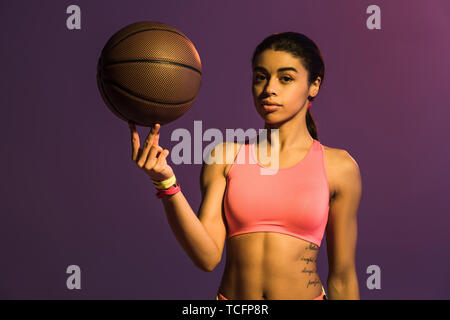 pretty african american girl in pink sports bra holding brown ball and looking at camera on purple background Stock Photo