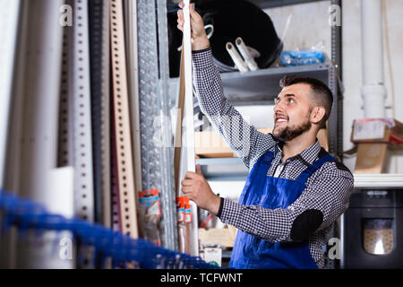 Young  smiling man worker examining plastic corners for tiles and floors in workshop Stock Photo
