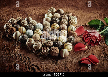 Heart of several artisan chocolates next to red roses with fallen petals sitting on top of cocoa powder Stock Photo