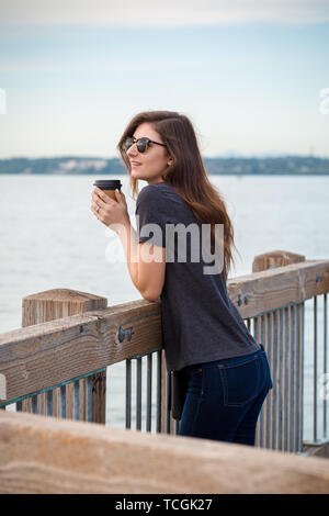 A pretty young brunette woman wearing sunglasses with a coffee on an oceanside boardwalk enjoying the ocean view. Stock Photo