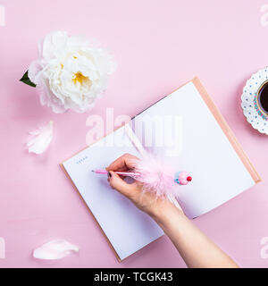 Woman is writing her Dreams in Notebook during drinking Morning Cup of Coffee. Pink background. Top View. Idea of Cozy Breakfast. Flat lay style. Stock Photo