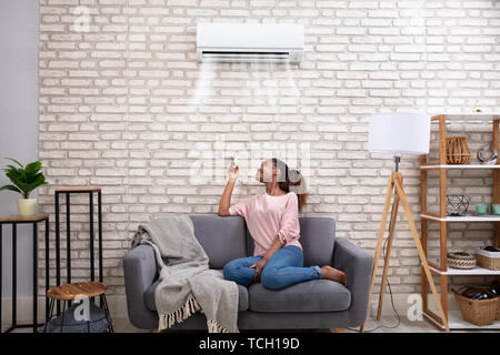 Young African Woman Switching On Air Conditioner With Remote Control At Home Stock Photo