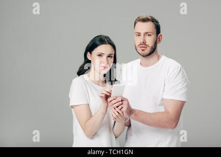 young man and woman in white t-shirts with usb cable around hands using smartphone isolated on grey Stock Photo