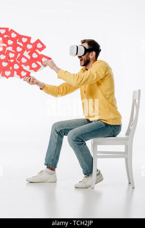 excited man in virtual reality headset sitting on chair and holding red paper cut cards with herts symbols on white background Stock Photo