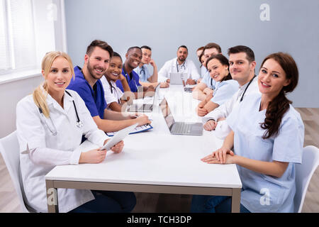 Happy Multi-ethnic Doctors Looking At Camera While Sitting At A Table In Medical Office Stock Photo