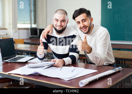 Two young smiling male students with thumbs up posing in classroom Stock Photo