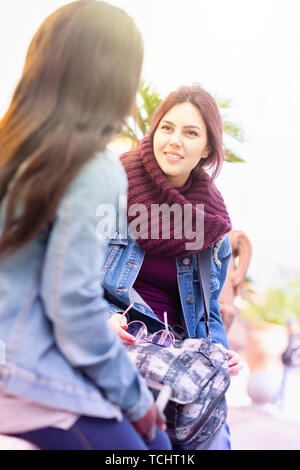 Two friends, of African and Caucasian ethnicity, talk sitting on a bench outdoors. Stock Photo