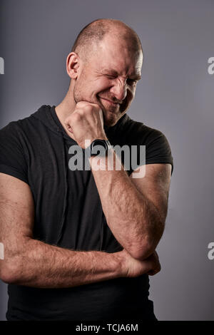 Thinking fun smiling man winking one eye and scratching the face in black casual shirt on grey background. Closeup portrait Stock Photo