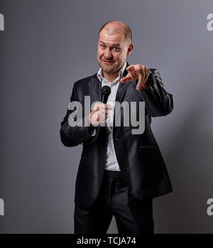 Fun emotional moving happy performer man presenting the show holding microphone in hand and showing the finger the choosing sign on grey color backgro Stock Photo