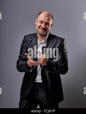 Fun emotional moving happy performer man presenting the show holding microphone in hand and showing the hand the welcome sign on grey color background Stock Photo