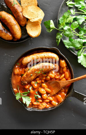 Grilled sausages with baked white beans in tomato sauce in frying pan over black stone background. Top view, flat lay Stock Photo