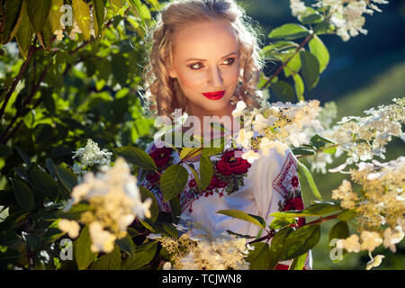 Closeup portrait of attractive blonde young woman with makeup and curly hairstyle in posing near bush with white flowers in garden. looking away and s Stock Photo