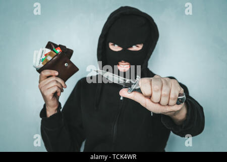 criminal robbery concept. An evil robber threatens with a knife and holds a stolen purse in his hand. Mortal danger. The malevolent thief smiles. Stock Photo