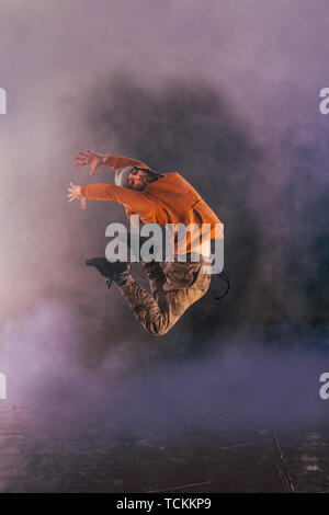 Male breakdancer jumps off high in the air while performing a cool dance move, a lot of smoke in the background. Stock Photo