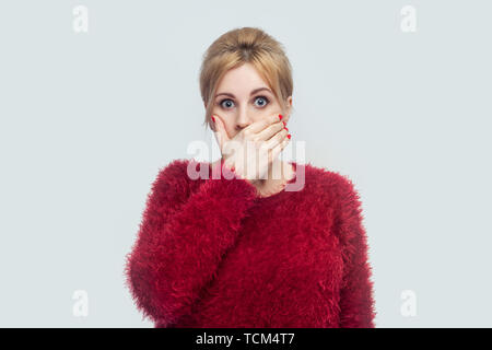 Portrait of shocked silence beautiful young blond woman in red blouse standing, covering her mouth and looking at camera with big eyes. indoor studio  Stock Photo