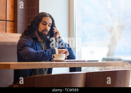 Portrait of serious worry handsome young adult man freelancer in casual style sitting in cafe, talking on the phone and looking to watch for planning  Stock Photo