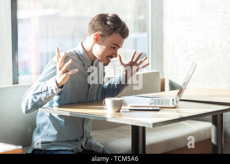 Portrait of aggressive unhappy young businessman in blue shirt are sitting in cafe and having bad mood are admonishing a worker through a webcam with  Stock Photo