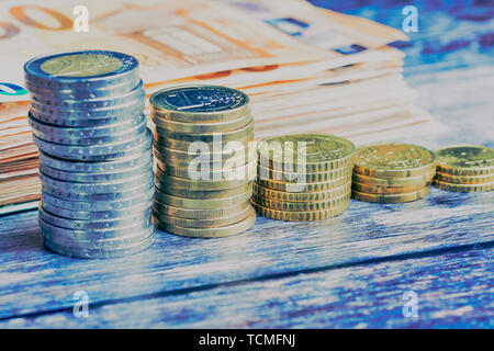 stacks of Euro coins and cent coins in front of blurred banknotes on a wooden background in a cold color style Stock Photo