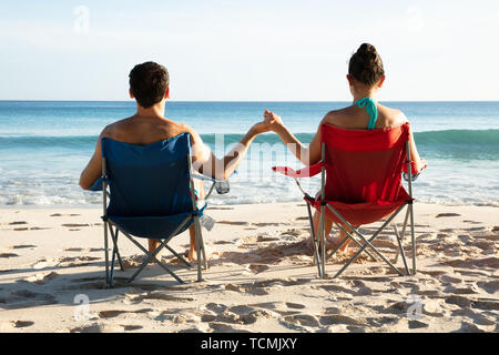 Rear View Of Young Couple Sitting On Chairs Holding Each Other's Hands Looking At Sea Stock Photo