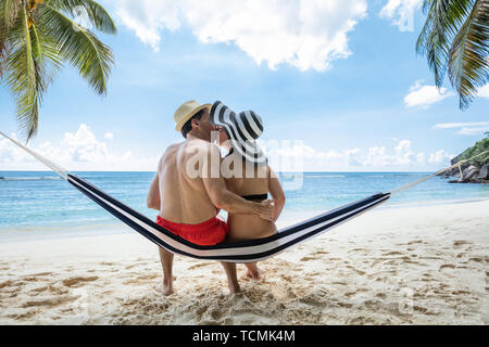 Rear View Of Young Couple Sitting On Hammock Kissing Near The Sea On Beach Stock Photo
