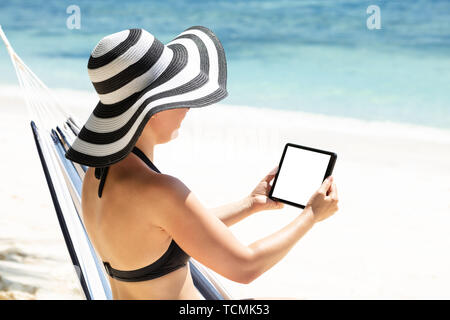 Rear View Of A Woman In Bikini Sitting On Hammock Using Digital Tablet At Beach Stock Photo