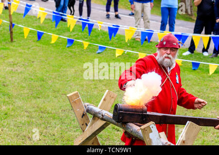 Le Medieval Weekend re-enactment event at Sandwich town in England. Man dressed in medieval red gunner outfit, firing a hand cannon mounted on a support. Smoke and fire erupting. Stock Photo