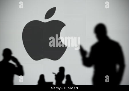 The Apple logo is seen on an LED screen in the background while a silhouetted person uses a smartphone in the foreground (Editorial use only) Stock Photo