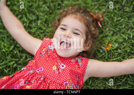 Happy Smiling Little Toddler Girl Laying On Grass In Park With Red Dress. Stock Photo