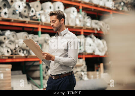 Manager going over his carpet inventory with a clipboard while standing on a warehouse floor with stacks of stock on shelves in the background Stock Photo