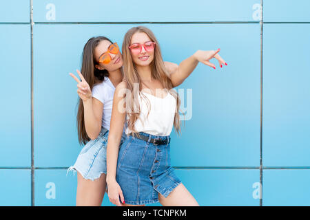 two young hipster woman friends in retro neon sunglasses standing and smiling over blue wall Stock Photo