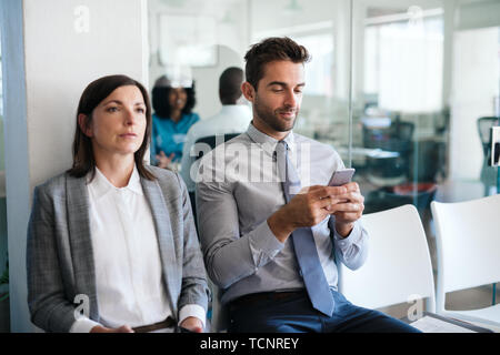 Smiling young man texting on his cellphone while sitting with another job applicant waiting for his interview Stock Photo