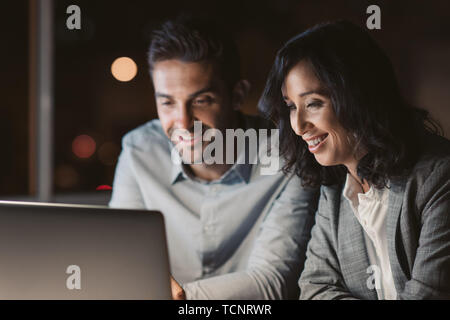 Two smiling businesspeople sitting at a desk using a laptop while working overtime together in an office at night Stock Photo