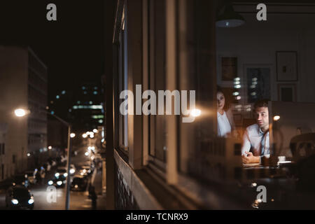 Two young colleagues working overtime together at a desk late at night behind office building windows reflecting the city Stock Photo